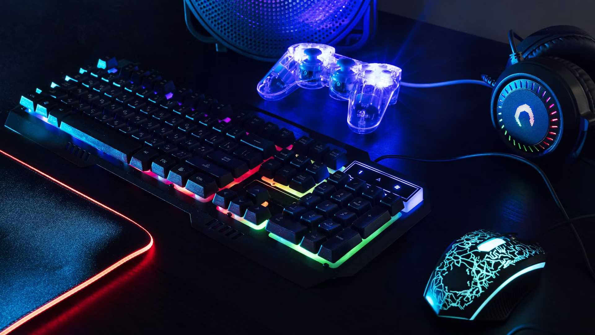 An illuminated RGB gaming keyboard, transparent controller, and mouse on a dark desk.