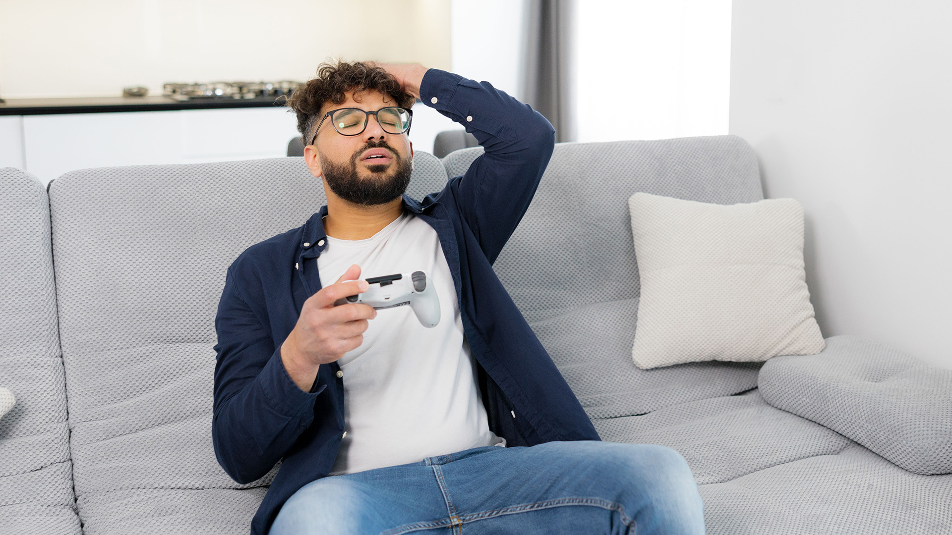 Frustrated man with glasses holding a gaming controller on a grey sofa.