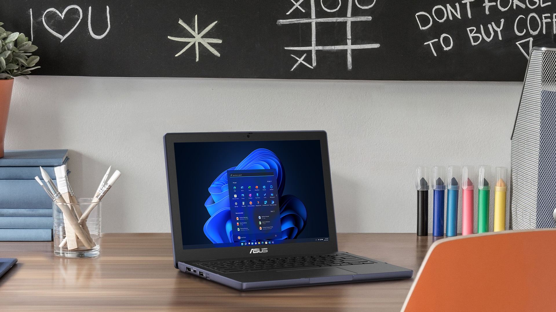 An ASUS Chromebook displaying Windows 11 on a desk next to art supplies.