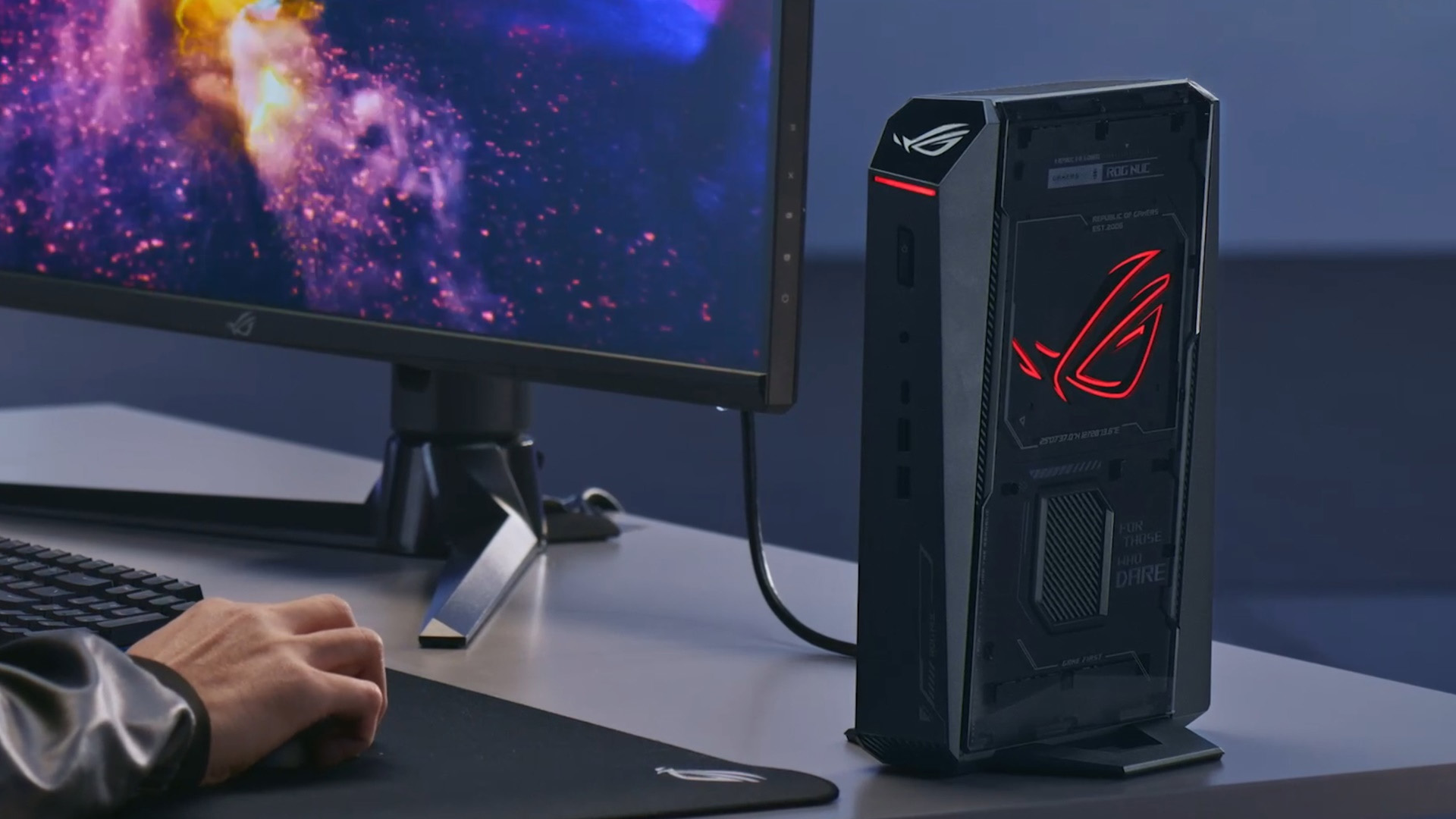 Compact ASUS ROG gaming desktop standing upright on a desk next to a monitor displaying a game.
