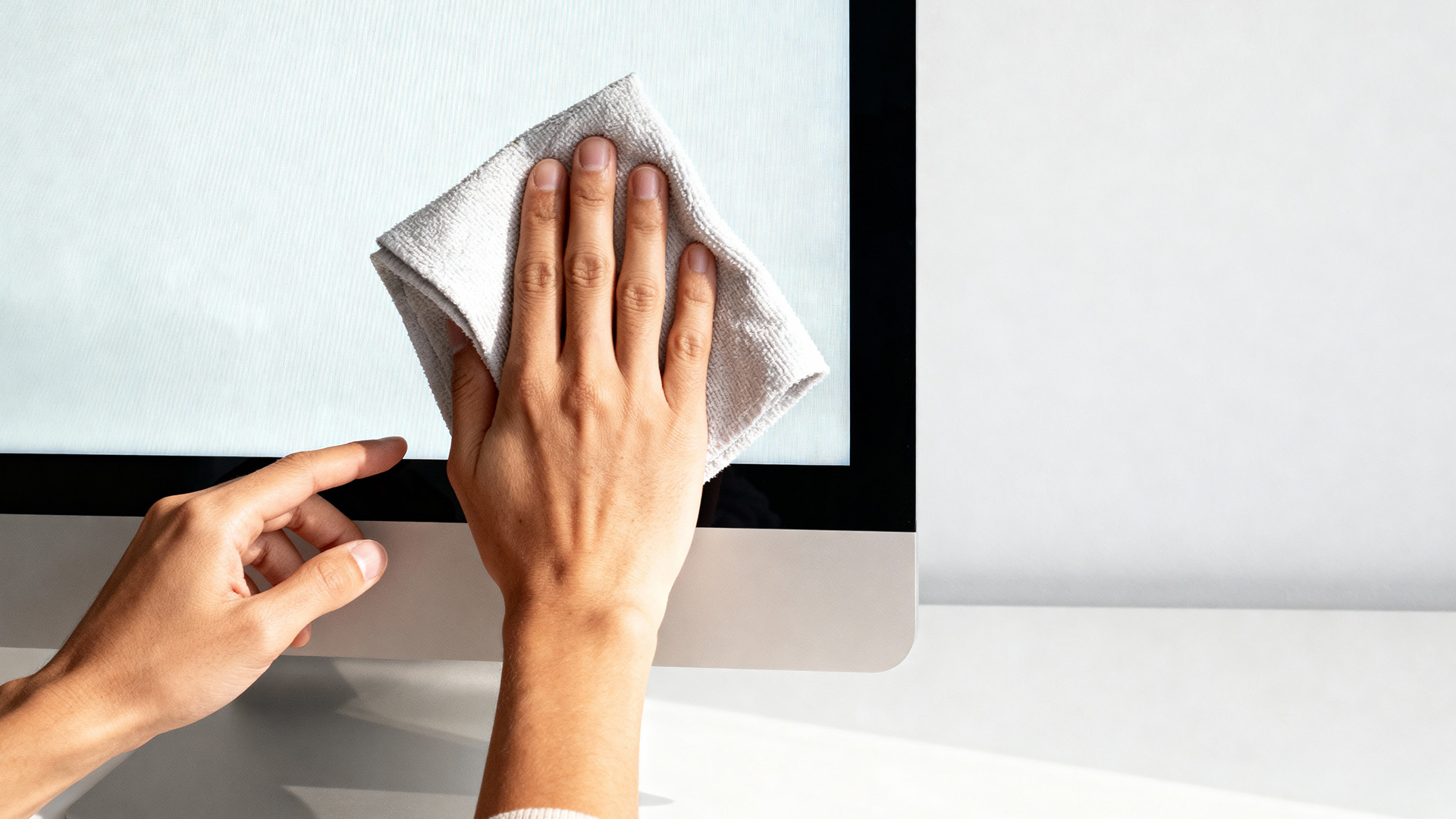 A close-up view of a hand wiping a computer screen with a microfibre cloth.