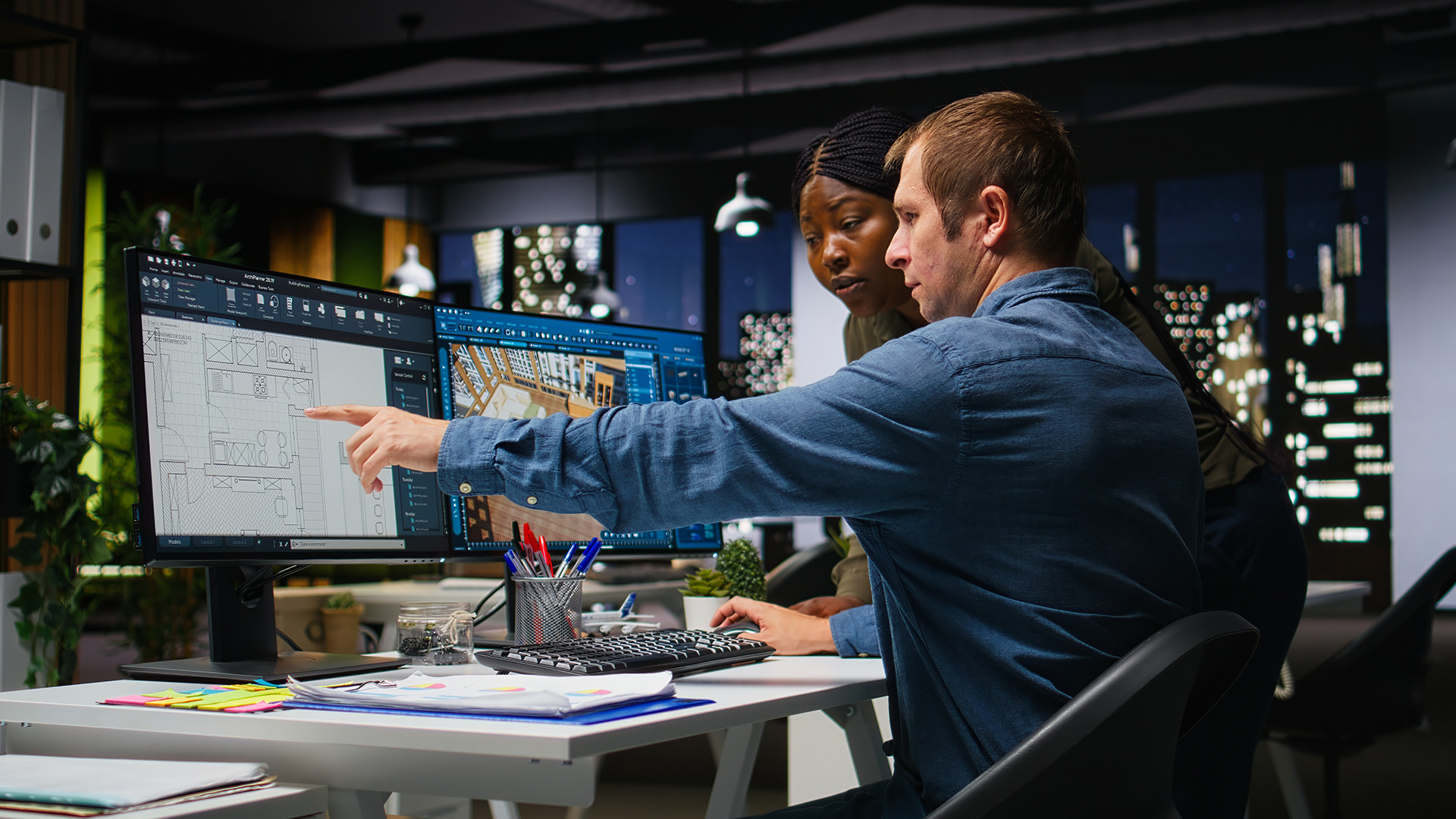 Two colleagues reviewing CAD architectural plans on a dual monitor setup in an office.