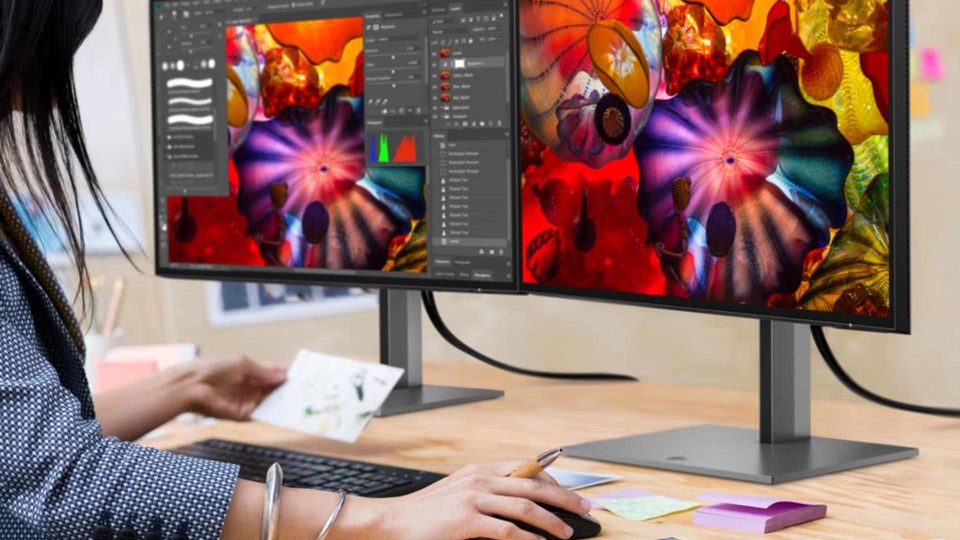 Woman editing colorful artwork on dual monitors at a wooden desk.