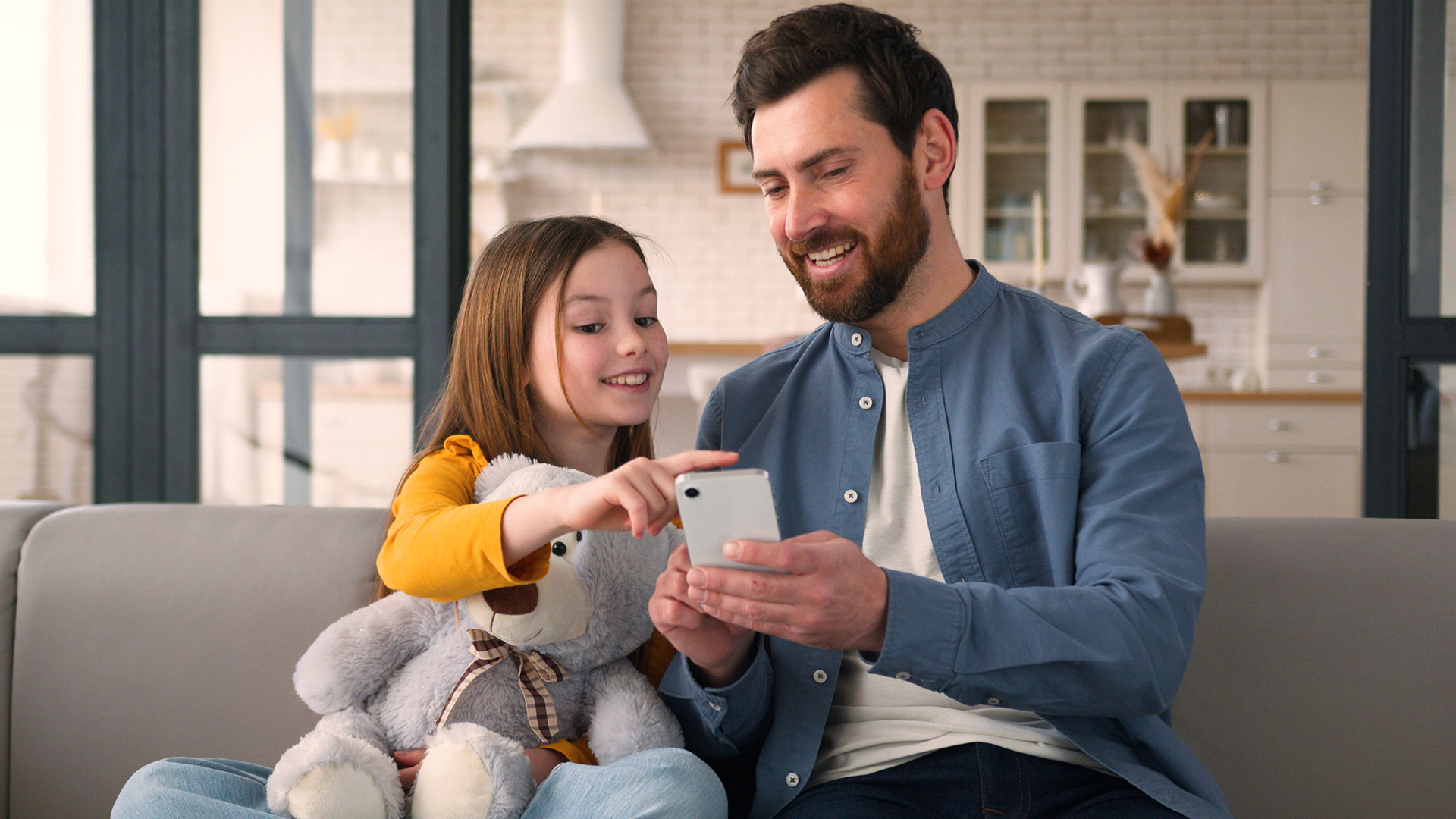 A smiling father and young daughter looking at a smartphone together on a sofa.