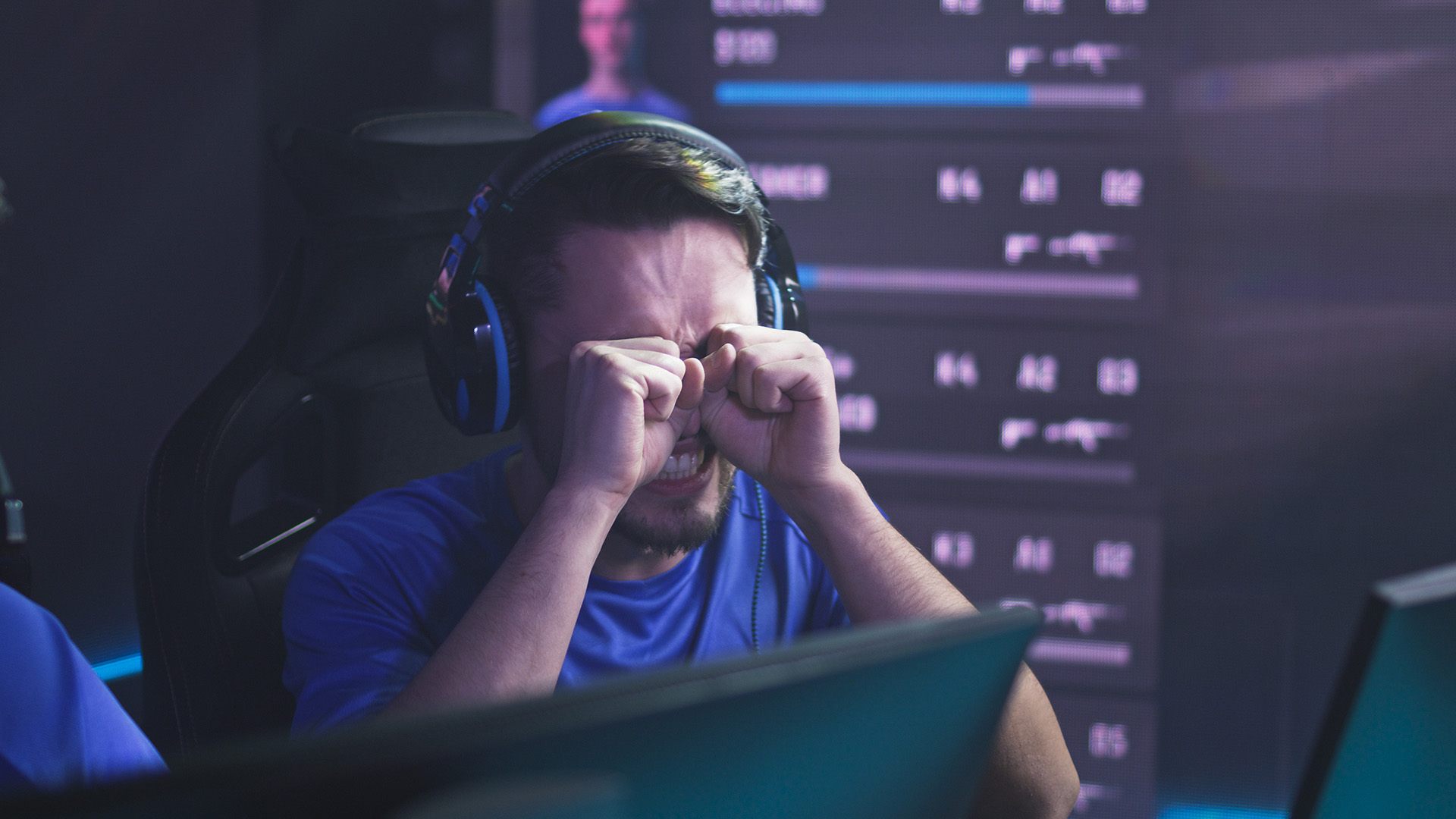 A frustrated gamer wearing a headset covering his eyes in front of monitors.