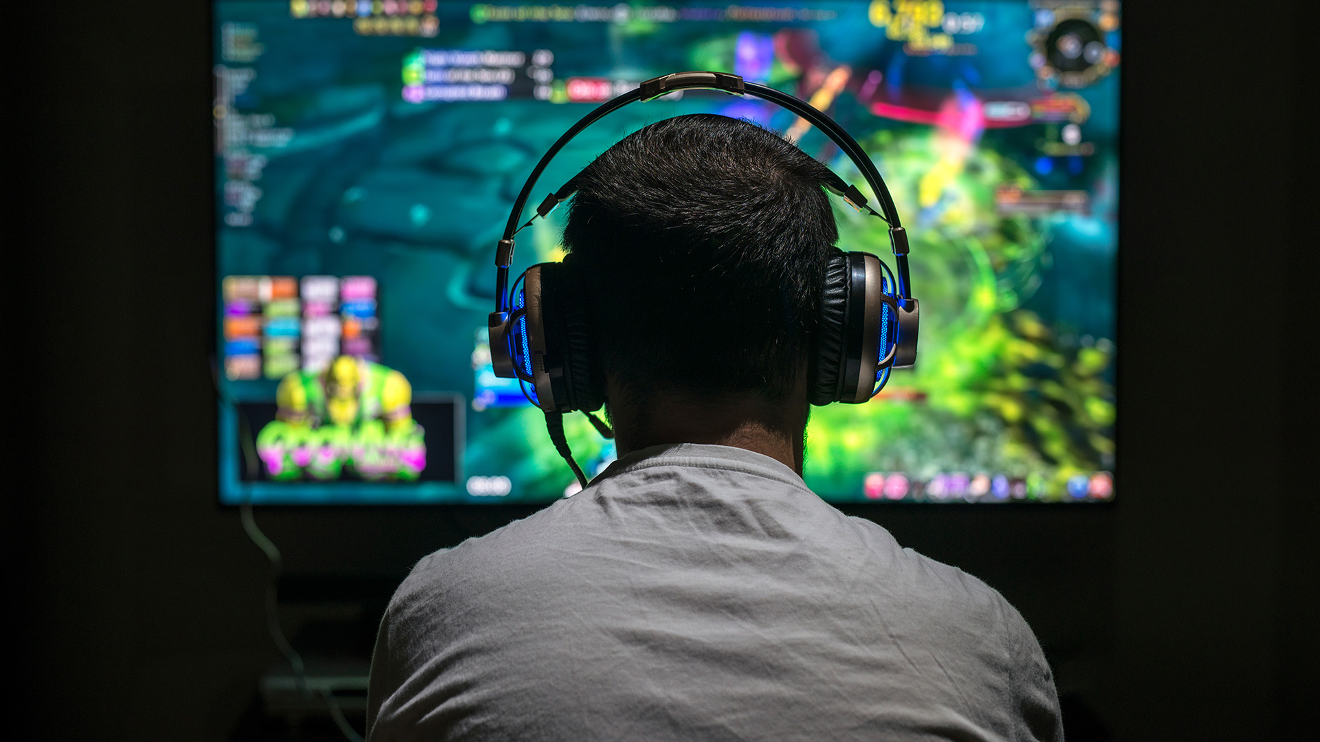 Rear view of a gamer wearing an illuminated headset playing a video game on a large screen.