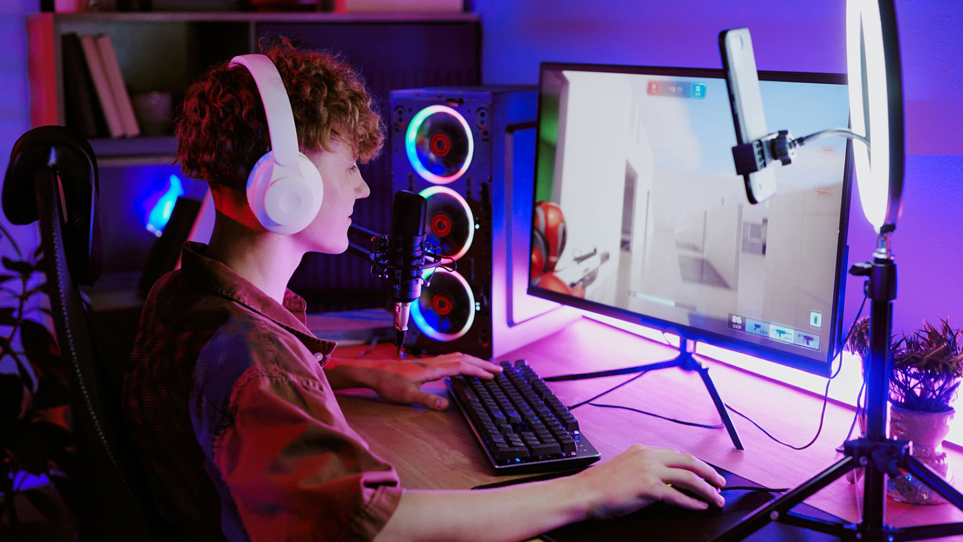 A young gamer wearing headphones playing a shooter game on an RGB gaming PC setup.
