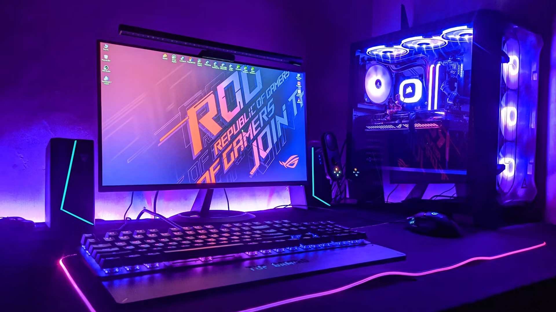 A gaming desk setup illuminated with purple lighting, featuring an ASUS ROG monitor and a custom PC case.