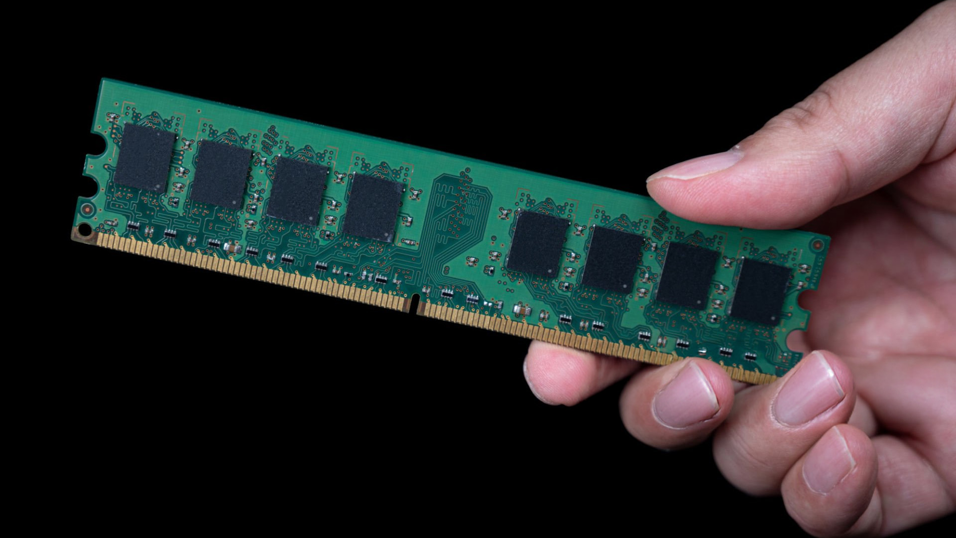 A person's hand holding a green desktop RAM stick against a dark background.