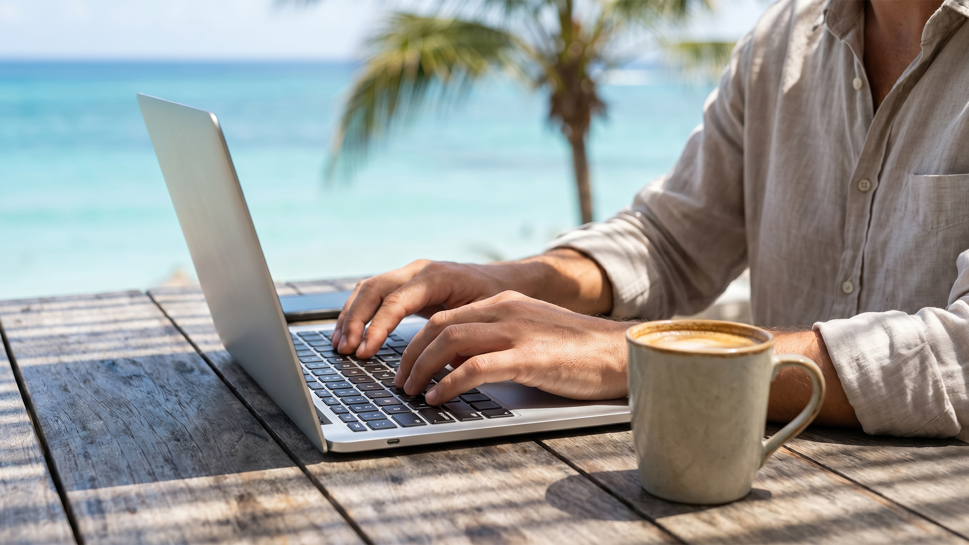 Close-up of hands typing on a laptop at a wooden table by the beach.