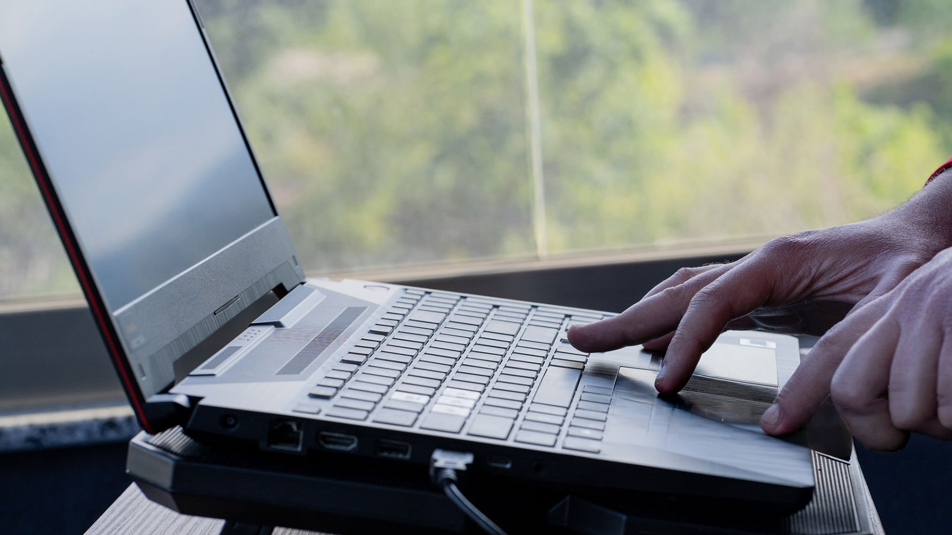 A person's hands typing on a laptop keyboard resting on a cooling stand near a window.