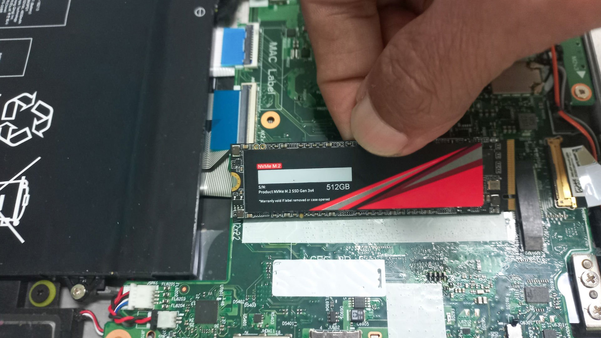A person's hand installing a 512GB NVMe M.2 SSD into a laptop motherboard slot.