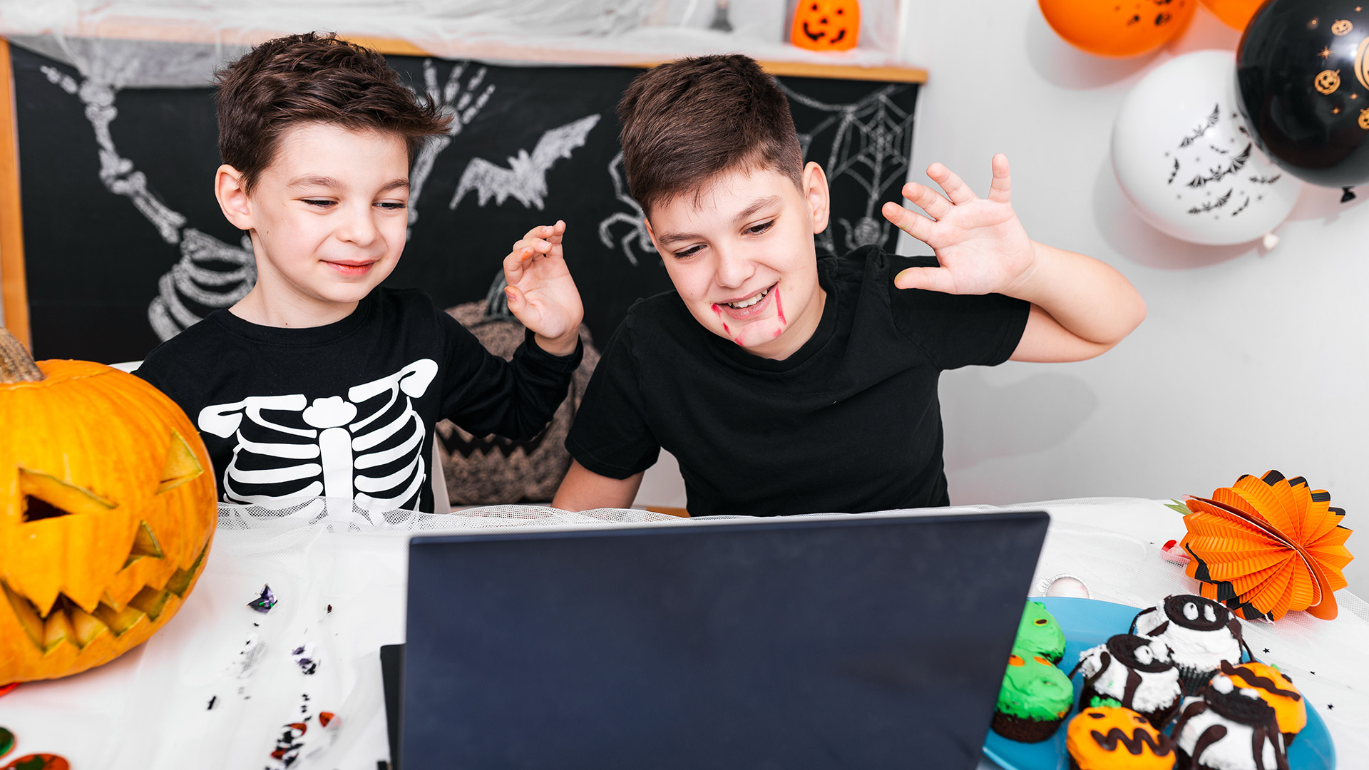 Two boys in Halloween costumes smiling and waving in front of a laptop.