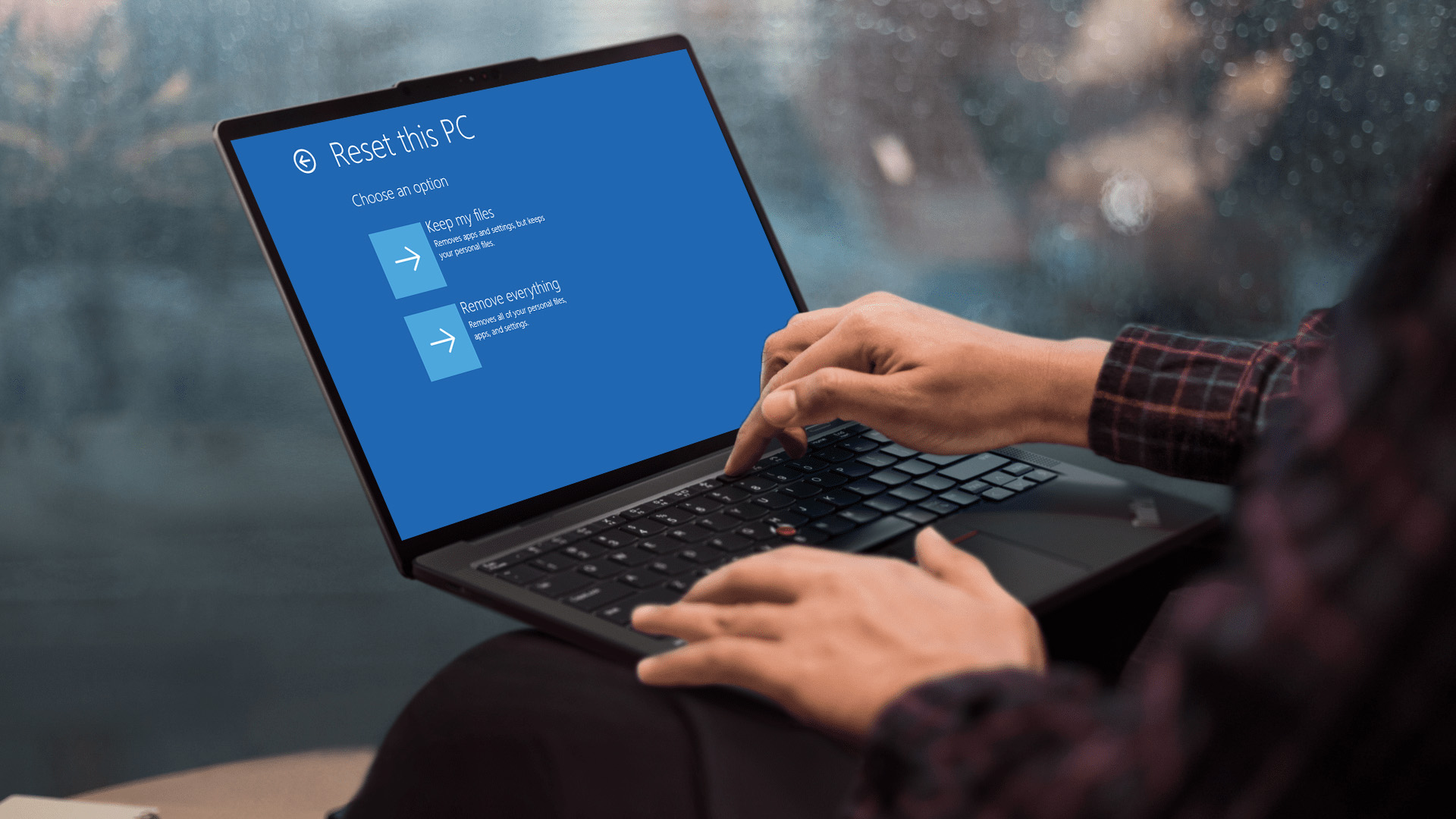 Hands typing on a Lenovo laptop showing the Windows Reset this PC screen.