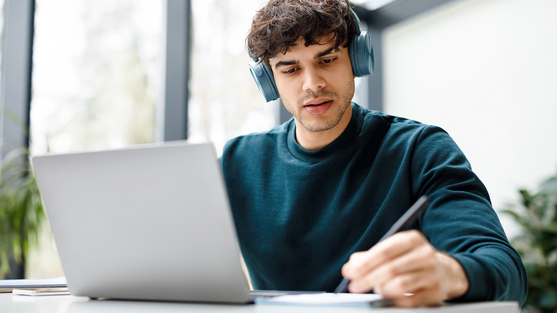 A male student wearing headphones while taking notes next to his laptop at a desk.