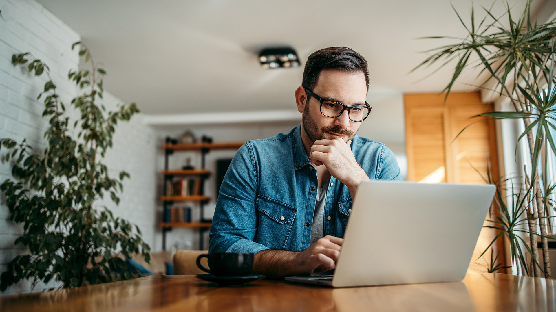 A man with glasses thoughtfully working on a laptop at a wooden desk.