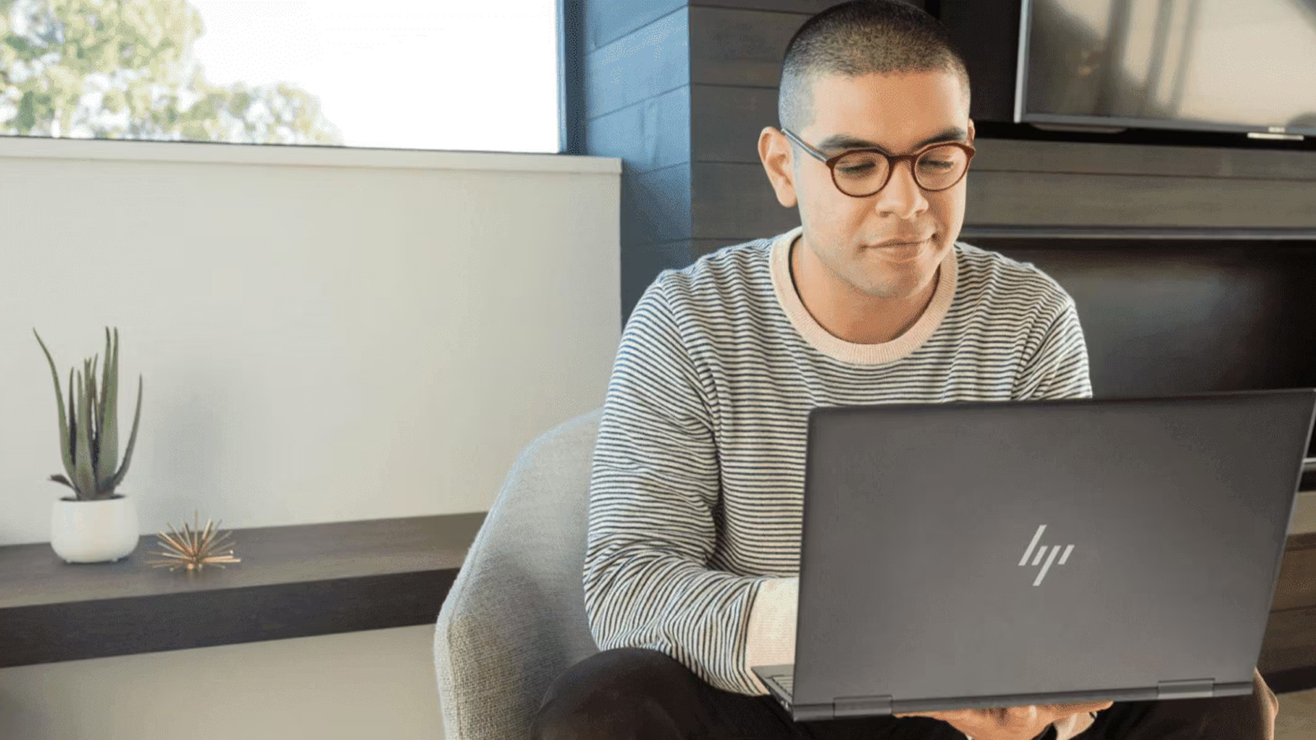 Man with glasses sitting and working on a dark HP laptop at home.