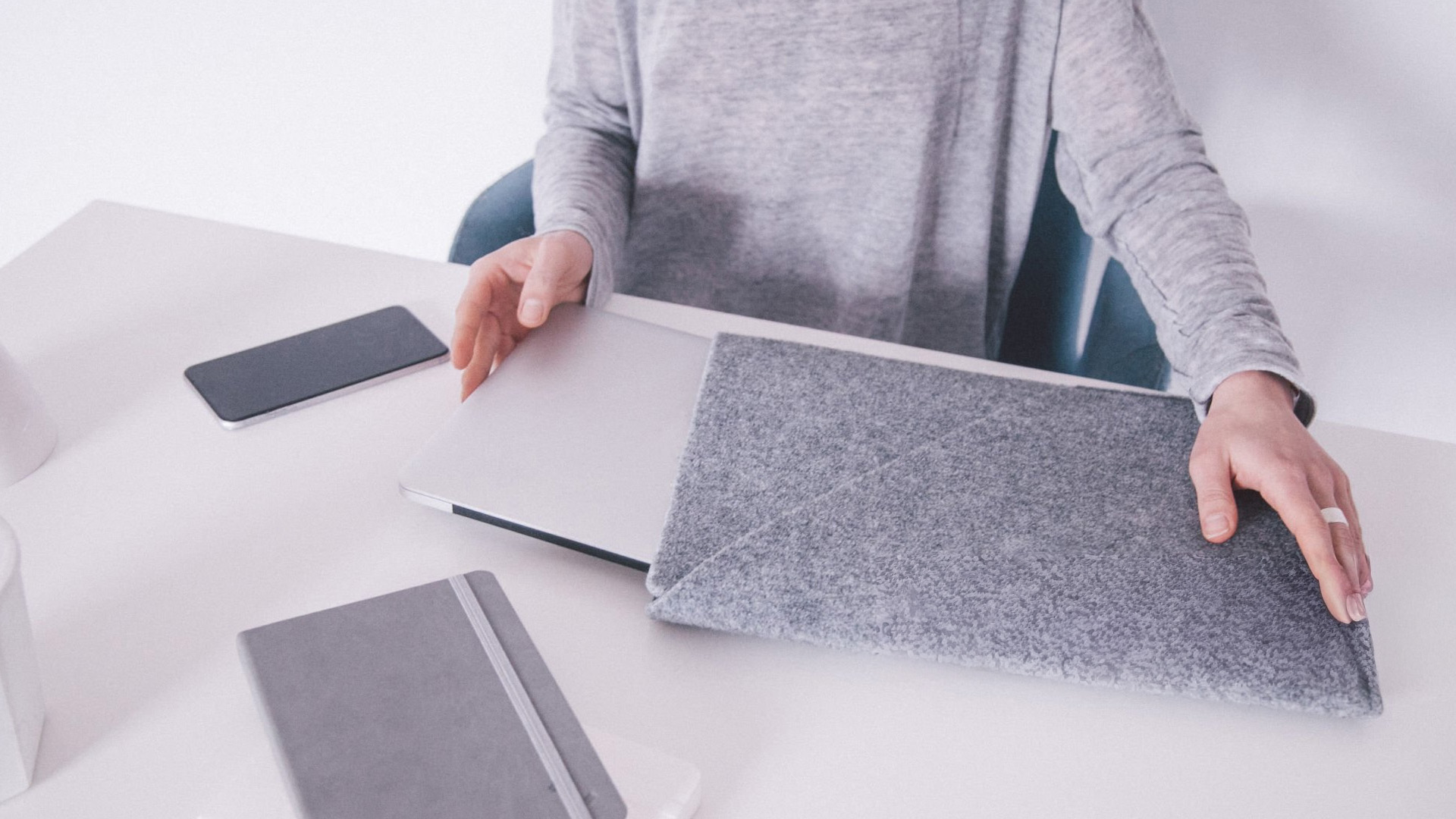 Person sliding a silver laptop into a grey felt protective sleeve at a desk.