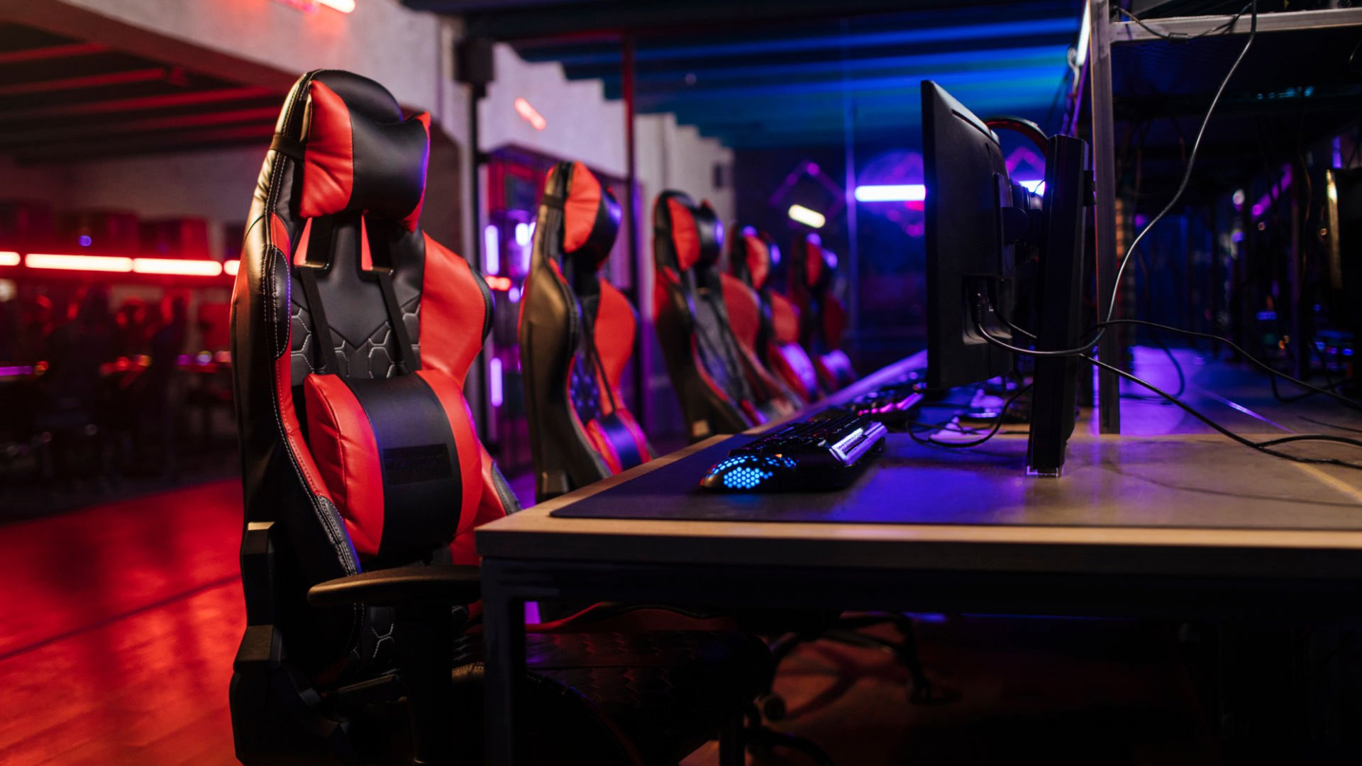Row of red and black gaming chairs at computer desks in a neon-lit room.
