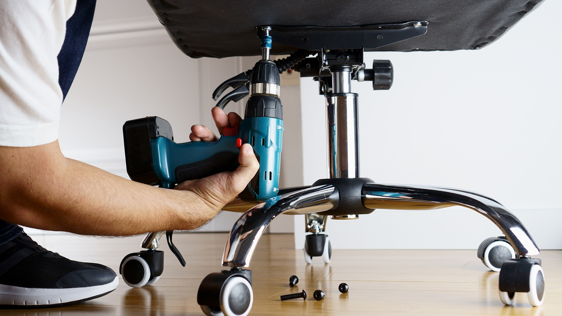 A person using a power drill to tighten bolts on the underside of a gaming chair.