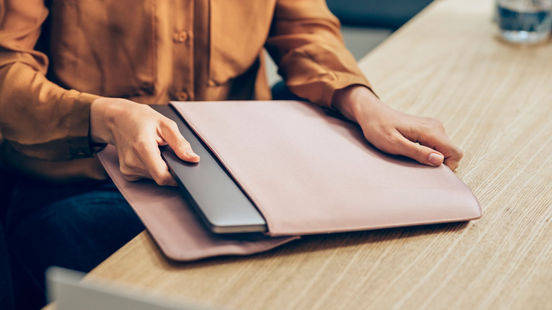 Hands placing a laptop into a pink leather sleeve on a wooden table.