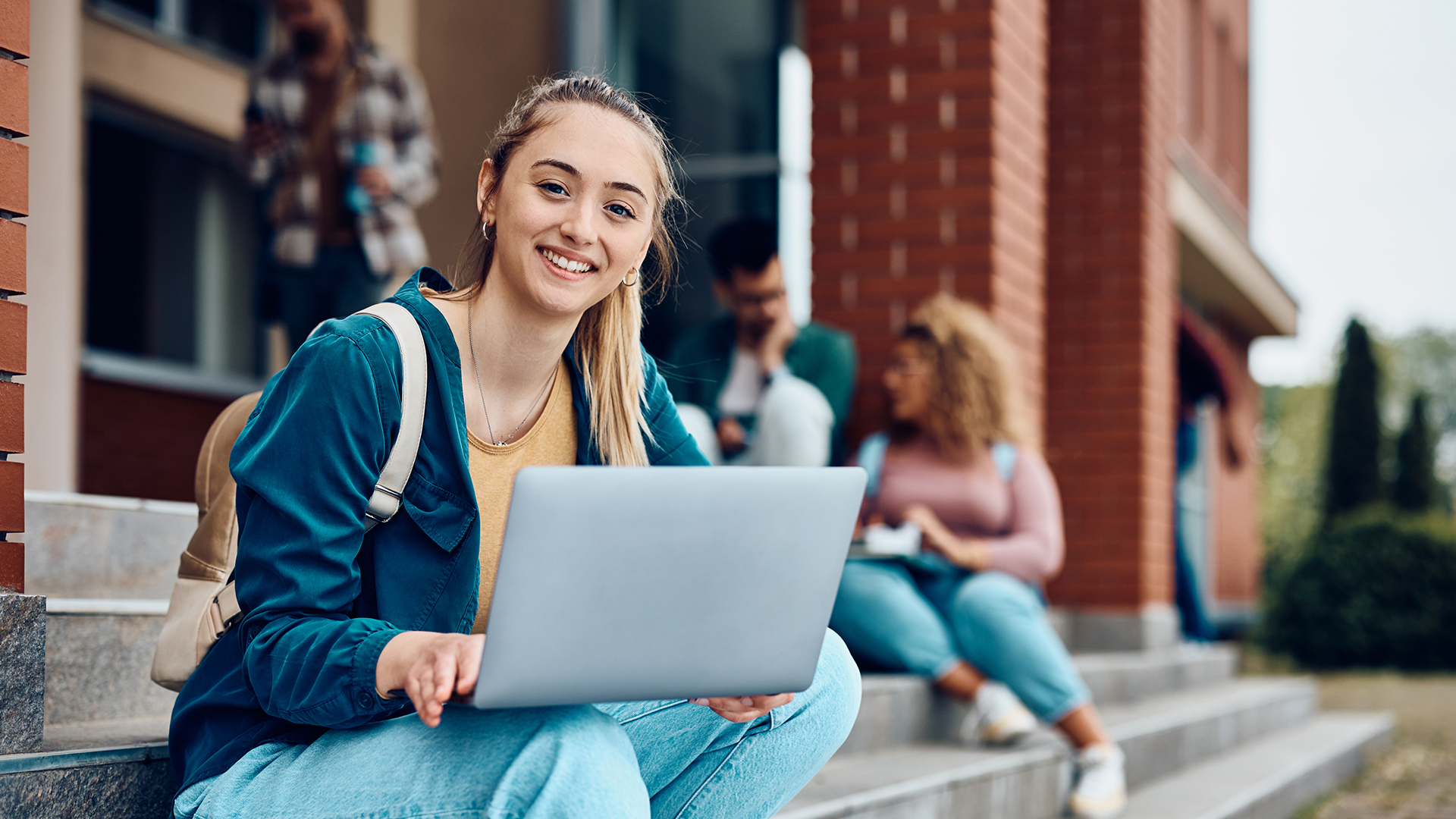 A smiling female university student holding a laptop while sitting on campus steps.