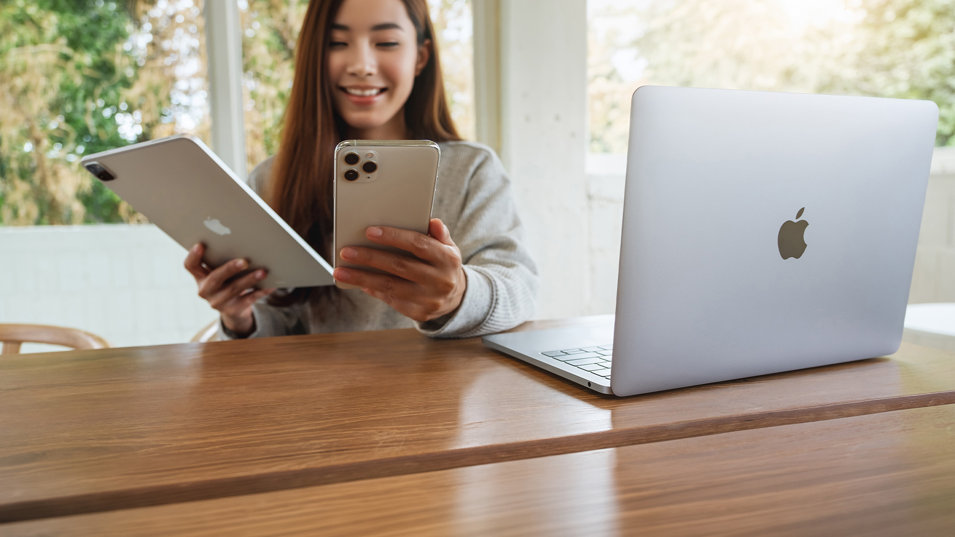 A smiling woman holding a tablet and smartphone next to a MacBook laptop.