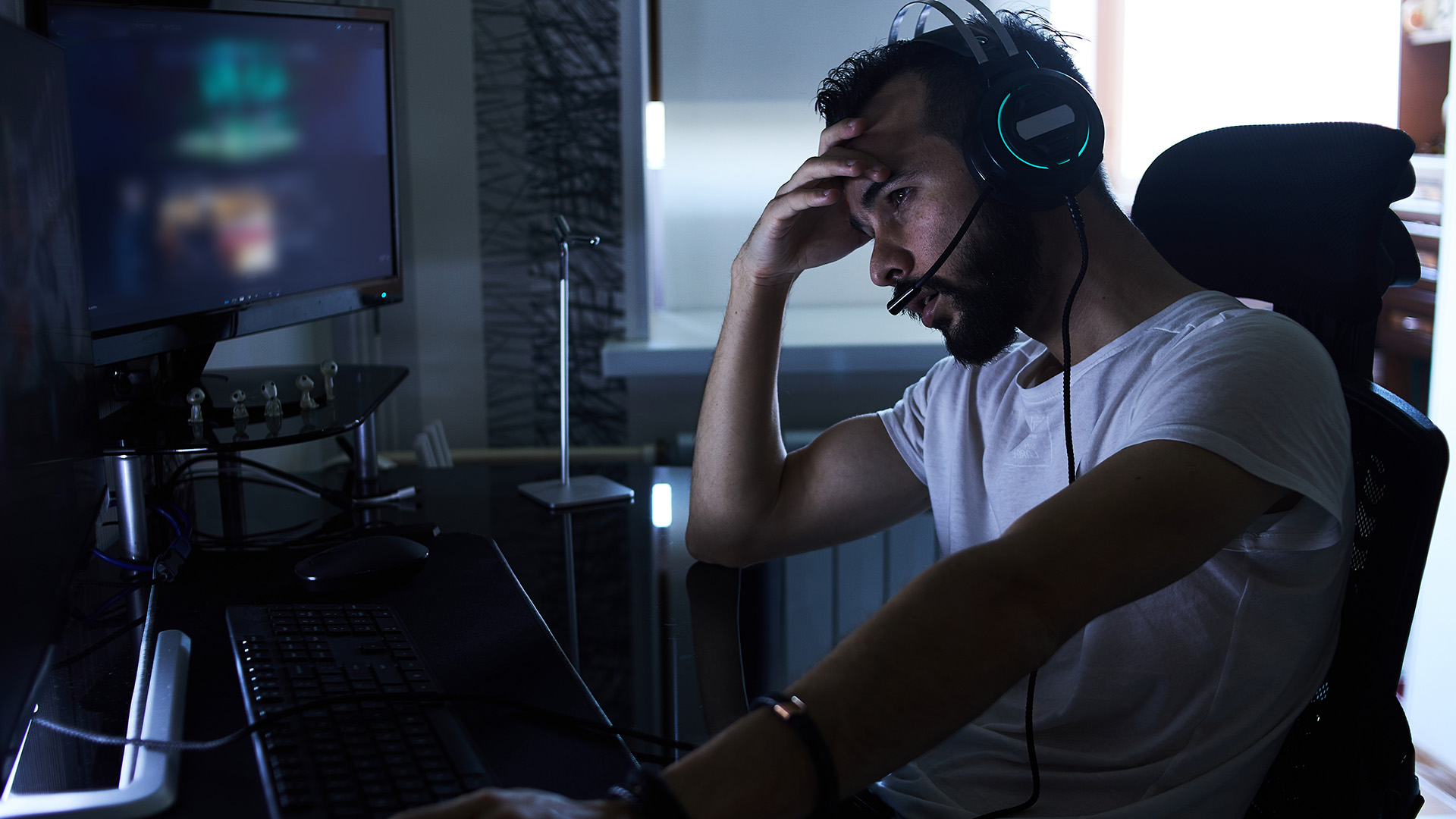 A stressed gamer wearing a headset sitting at a desk with computer monitors.