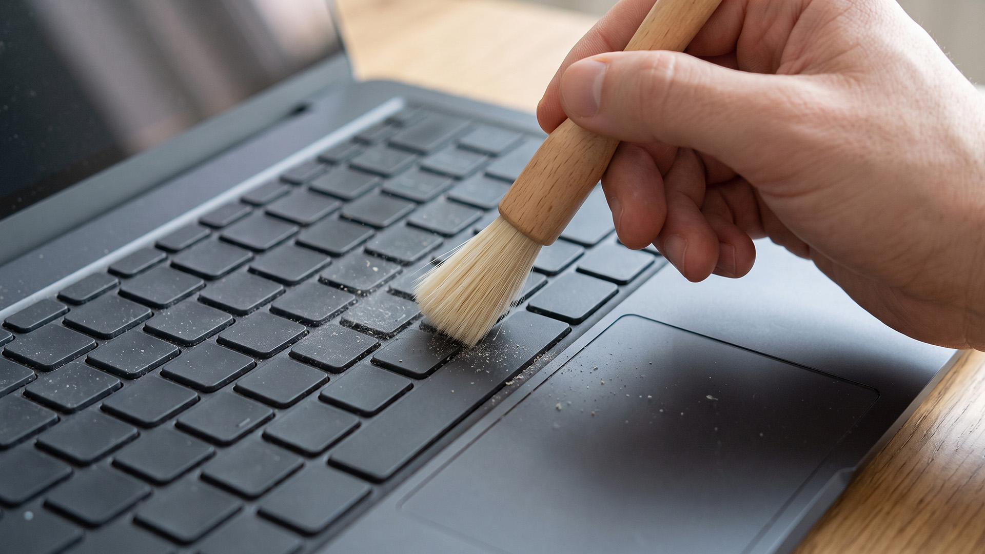 Hand using a soft bristle brush to sweep dust off a dark laptop keyboard.