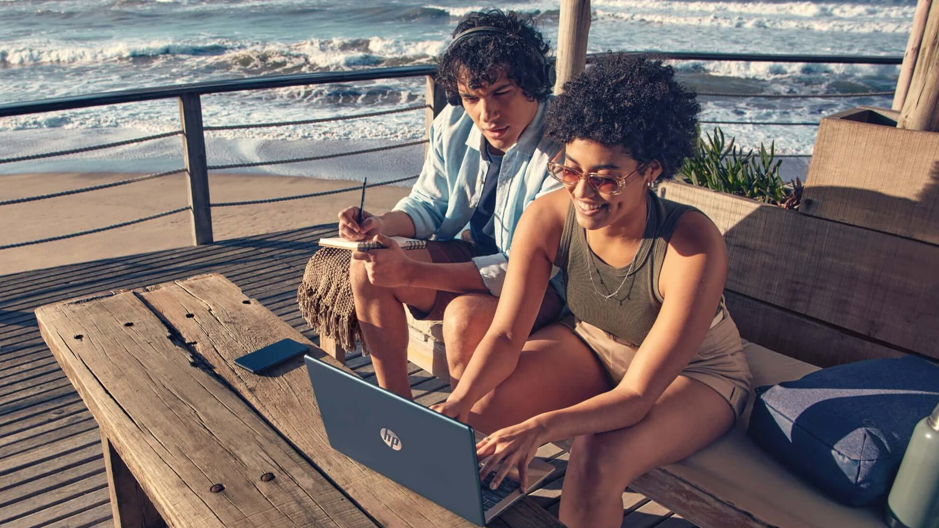 Two people using a blue HP laptop outdoors near the beach.