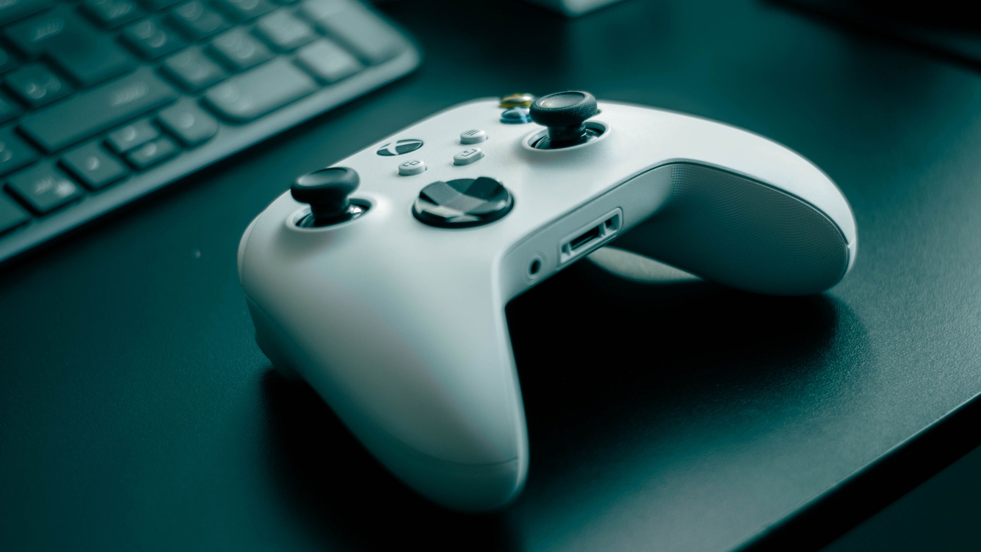A white Xbox wireless controller resting on a dark desk beside a keyboard.