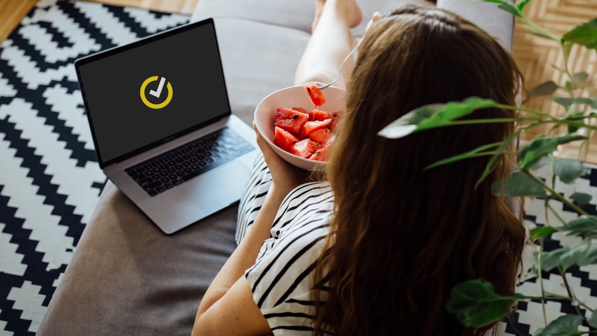 A woman eating watermelon while looking at a laptop displaying the Norton checkmark logo.