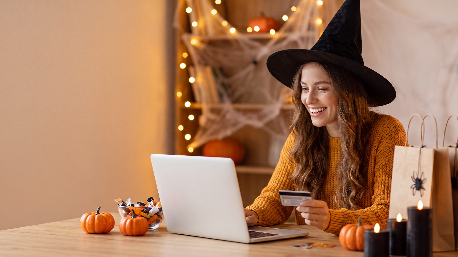 A smiling woman wearing a witch hat holding a credit card while using a laptop.