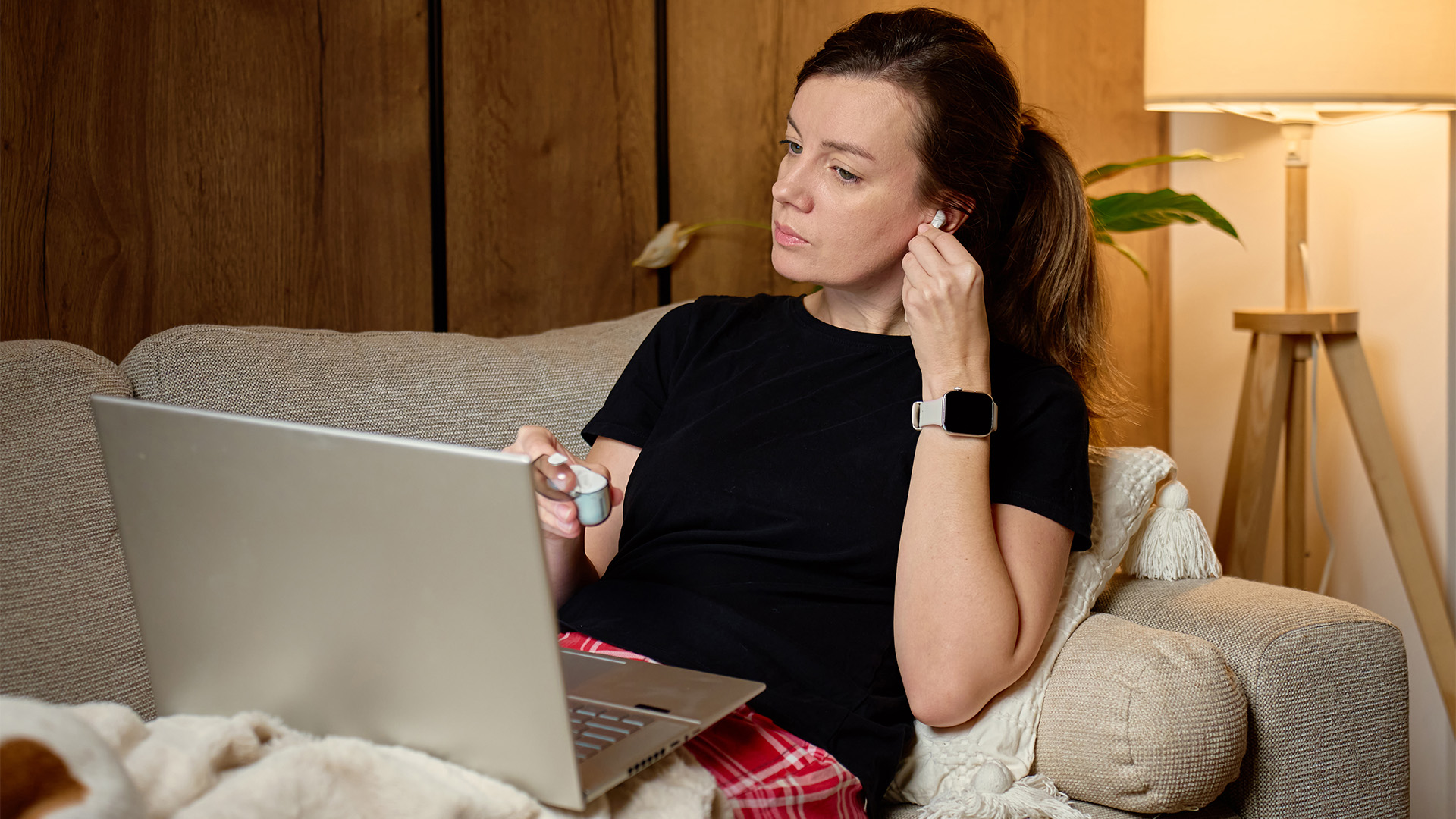 A woman sitting on a couch wearing wireless earbuds while using her laptop.