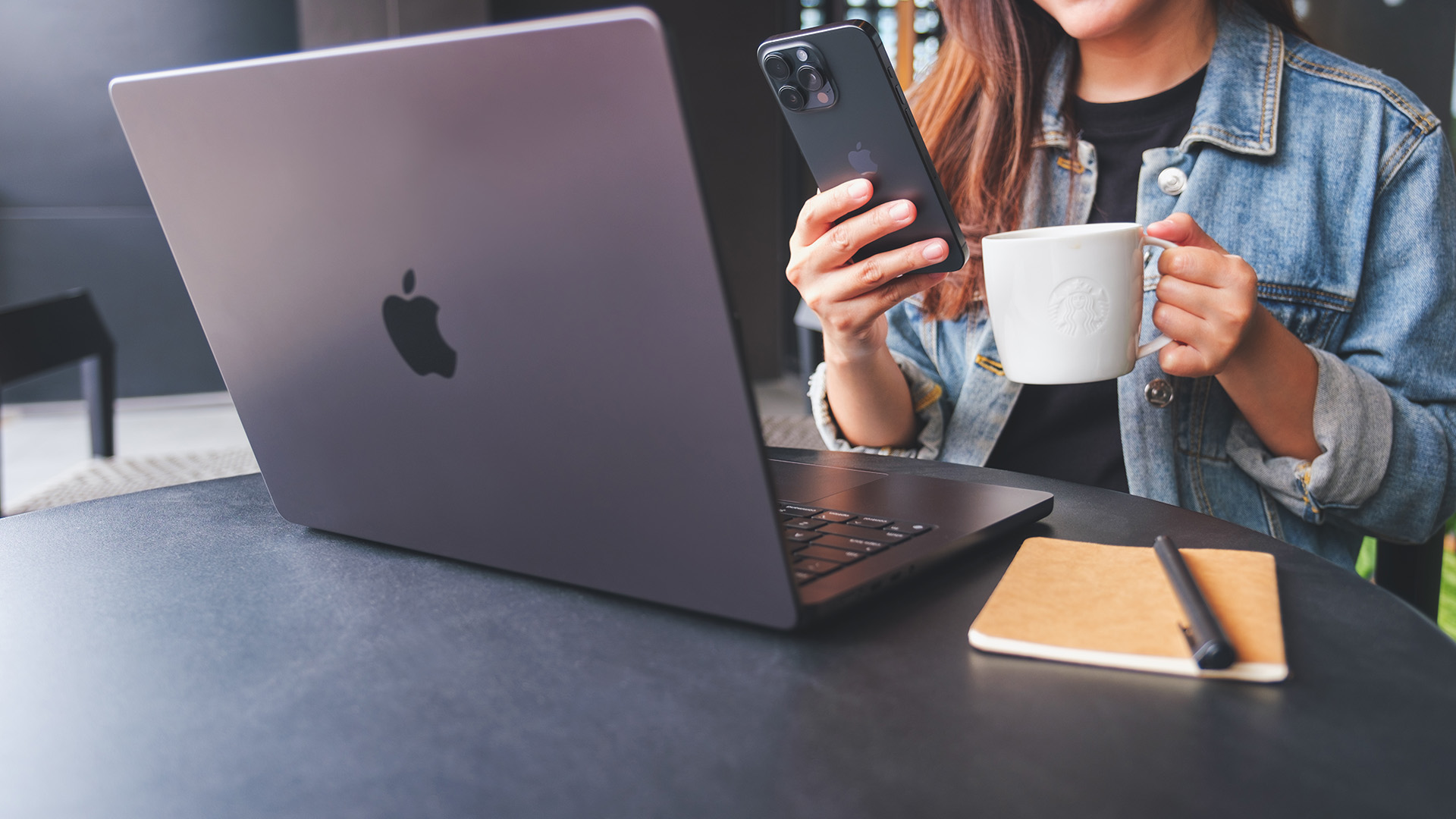 A woman using an Apple MacBook laptop and holding an iPhone at a cafe table.