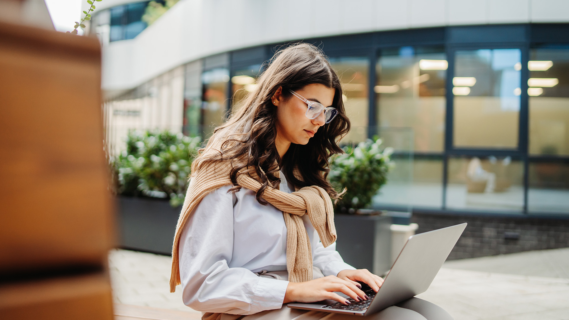 A woman wearing glasses typing on a laptop outdoors in a modern courtyard area.