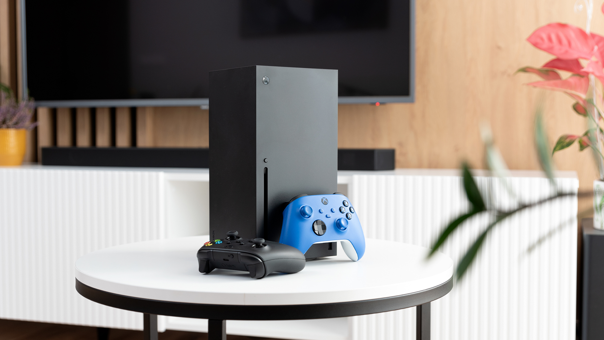 An Xbox Series X console set up on a white table with controllers in front of a large television screen.