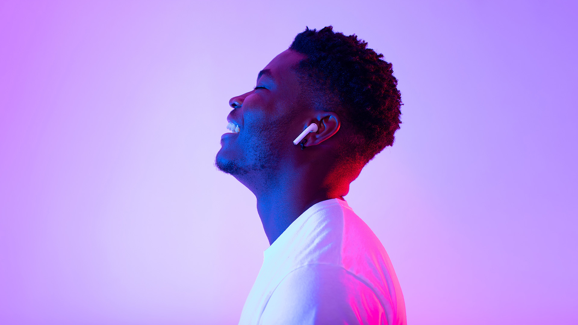 Young man enjoying music with wireless earbuds in purple lighting.
